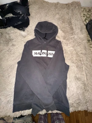 Svart hoodie från Madness - Säljer en svart hoodie från Madness med stor vit logga på bröstet och en liten patch på fickan. Klassisk känguruficka och justerbar huva med snören. Perfekt för dig som gillar streetwear och vill ha en enkel men snygg look.