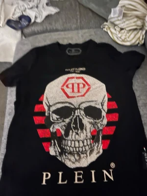 Svart t-shirt från Philipp Plein - Cool svart t-shirt från Philipp Plein med stort tryck framtill i rött, vitt och silver. T-shirten har rund hals och korta ärmar. Perfekt för dig som gillar statement-plagg och streetstyle.