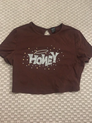 Brun croppad t-shirt med tryck - Snygg brun kort croppad t-shirt  med texten 'Honey' i vitt och guldprickar på framsidan. T-shirten har korta ärmar och rund halsringning. Perfekt för en trendig och avslappnad stil.