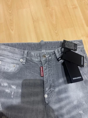 Grå slim jeans från Dsquared2 - Snygga grå jeans från Dsquared2 med slitna detaljer och smal passform. Jeansen har klassisk femficksdesign, silverfärgade knappar och en röd logotagg vid gylfen. Perfekta för en trendig och avslappnad stil.