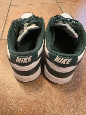 Nike Dunk Low grön/vit sneakers - Snygga Nike Dunk Low sneakers i grönt och vitt läder med klassisk Nike-logga på hälen och sidan. Skorna har gröna skosnören och perforerad tå för extra ventilation. Perfekta för dig som gillar streetwear och vill ha en stilren look.