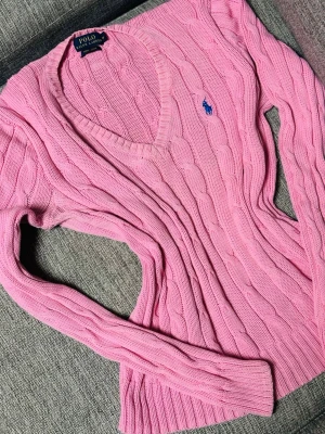 Rosa kabelstickad tröja Polo Ralph Lauren - Snygg rosa kabelstickad tröja från Polo Ralph Lauren i mjuk pima cotton. Tröjan har v-ringning, långa ärmar och den klassiska blå broderade loggan på bröstet. Perfekt för dig som gillar preppy stil och vill ha något bekvämt och trendigt.båda två för 1300