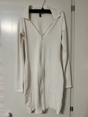Klänning - Vit ribbad kortklänning från H&M med lång ärm, krage och dragkedja framtill. Klänningen har en figurnära passform och är tillverkad i ett stretchigt material. Perfekt för en trendig och stilren look. Aldrig använd.