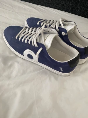 Oliver cabell Yale - Snygga blå sneakers från Oliver Cabell med vita detaljer och vita skosnören. Skorna har en låg profil, svart hälparti och en vit sula. Tillverkade i slätt läder med en stor vit cirkeldetalj på sidan. Perfekta för en clean och modern stil.