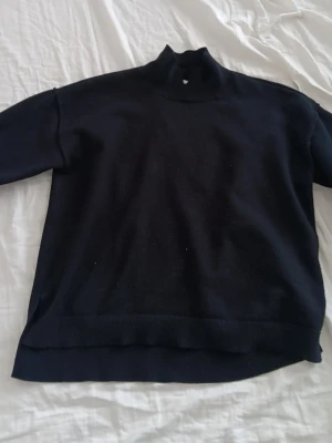 Svart stickad polotröja - En svart stickad polotröja med lång ärm och ribbade muddar vid ärmslut och nederkant. Tröjan har en hög krage och en rak, avslappnad passform. Perfekt för lager-på-lager och enkel att matcha med olika outfits.