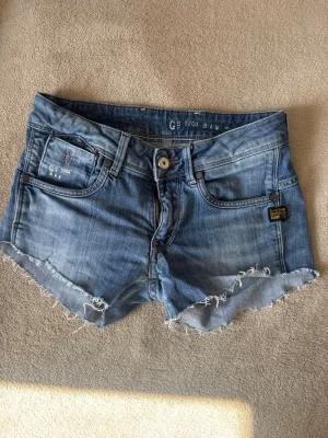 Blå jeansshorts G-Star - Säljer ett par blå jeansshorts från G-Star med råa, fransiga kanter och klassisk femficksdesign. Shortsen har normal passform och är tillverkade i bomull. Perfekta för sommaren och har en avslappnad streetstyle vibe.