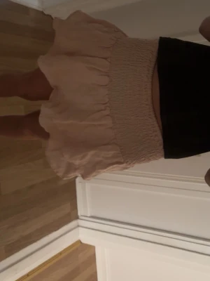 Rosa volangkjol med smock - Söt kort kjol i rosa med smock i midjan och volang nertill. Kjolen har en luftig och lätt känsla, perfekt för varmare dagar. Passar dig som gillar en feminin och trendig stil. Kjolen är från barnavdelningen och är i storlek 13-14.