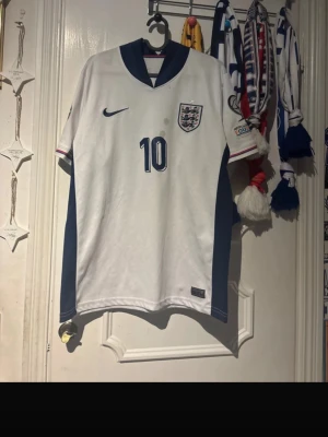 England fotbollströja Nike #10 - Säljer en vit England fotbollströja från Nike med nummer 10 på bröstet och officiellt landslagsmärke. Tröjan har blå detaljer längs sidorna och kragen samt ett färgglatt EM-märke på ärmen. Perfekt för dig som älskar fotboll och vill stötta England. Fläckar på som inte går bort