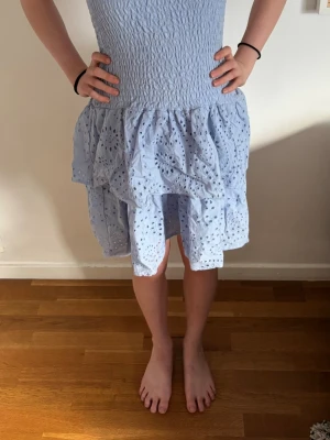 Ljusblå volangklänning med broderi - Söt ljusblå kortklänning med smock upptill och volanger nertill. Klänningen har tunna axelband och är dekorerad med broderade hålmönster. Perfekt för sommarens alla avslutningar och kalas! Storlek barn 146/152 men som XXS.