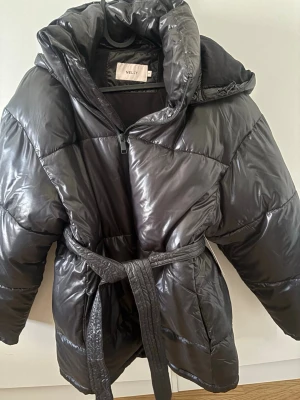 Svart pufferjacka från Nelly med bälte - Snygg svart pufferjacka från Nelly med glansig finish, stor huva och bälte i midjan för extra stilpoäng. Jackan har dragkedja framtill och är perfekt för kalla vinterdagar. Materialet är syntetiskt och jackan har en oversized look.