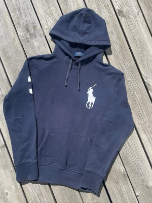 Mörkblå hoodie från Polo Ralph Lauren - Snygg mörkblå hoodie från Polo Ralph Lauren med stor vit polospelare-logga på bröstet och klassisk känguruficka. Huva med dragsko och ribbade muddar. Perfekt för en chill och stilren look. 