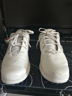 Golf skor från Under Armour - Snygga vita sneakers från Under Armour med lågt skaft och mörkblå detalj vid hälen. Skorna har klassisk snörning och ovandel i syntetmaterial med meshpartier för extra andningsförmåga. Perfekta för en sportig och avslappnad stil.