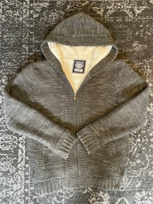 Grå stickad hoodie med teddyfoder - Mysig grå stickad hoodie med dragkedja och vit teddyfoder på insidan. Tröjan har två fickor framtill, ribbade muddar och en stor huva. Perfekt nu till våren eller sommarkvällar.