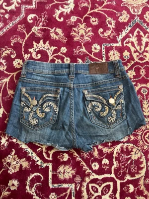 Blå jeansshorts med broderade fickor - Snygga blå jeansshorts från Texas med slitna kanter och detaljerade broderier på bakfickorna. Shortsens fickor har coola mönster i beige och vitt med nitar och knappar. Perfekta för en avslappnad och trendig look.