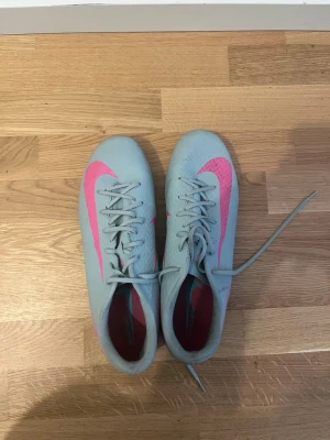 Nike Zoom Vapor 16  - Snygga fotbollsskor från Nike i ljusblått med en stor rosa swoosh på sidan. Skorna har snörning och låg profil, perfekt för snabba rörelser på planen. Yttermaterialet är syntetiskt och designen är modern och sportig.