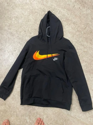 Svart Nike hoodie med färgglad logo - Svart hoodie från Nike med stor färgglad swoosh-logga i orange och gult på bröstet samt en mindre vit Nike-text. Klassisk känguruficka framtill och justerbar huva med dragsko. Perfekt för en sportig och avslappnad stil.