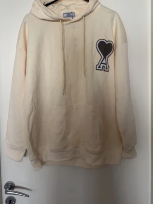 Beige Ami Paris hoodie med hjärtdetalj - Snygg beige hoodie från Ami Paris med stor svart hjärtdetalj och bokstaven A på bröstet. Hoodien har huva med dragsko och en stor magficka. Perfekt för en chill och trendig look. Mjuk bomullskänsla och avslappnad passform.