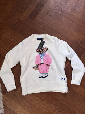 Vit stickad tröja med Polo Bear - Vit stickad tröja från Polo Ralph Lauren med ikoniska Polo Bear-motivet framtill. Björnen har på sig en rosa kostym och håller en blomma. Tröjan har rund halsringning, ribbade muddar och RL-broderi nertill. Perfekt för dig som gillar preppy och lekfull stil.