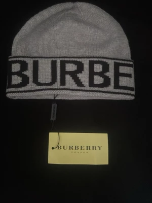 Grå Burberry mössa - Snygg grå mössa som har märket Burberry med bred svart logotyp runt hela kanten. Klassisk beanie-modell i stickat material som håller dig varm och ger en trendig look. Perfekt accessoar för att lyfta din outfit med en lyxig touch. Helt ny och jätte snygg 
