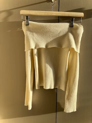 Ribbad offshoulder topp i beige - Trendig ribbad offshoulder topp i beige med lång ärm. Toppen har en croppad passform och är tillverkad i ett mjukt, stretchigt material som sitter snyggt på kroppen. Perfekt för dig som vill ha en stilren och modern look.