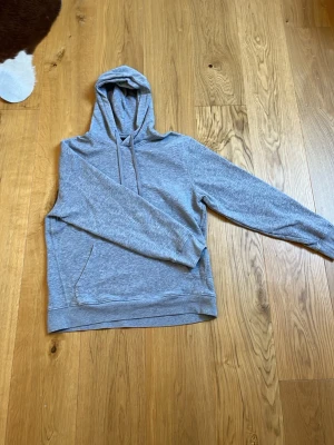 Ljusgrå basic hoodie med ficka - En stilren ljusgrå hoodie med huva och dragsnören. Hoodien har en klassisk känguruficka framtill och ribbade muddar vid ärmslut och nederkant. Perfekt för en avslappnad look. Materialet känns mjukt och bekvämt mot huden.