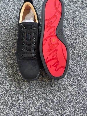 Svarta Louboutin sneakers med röd sula - Snygga svarta sneakers från Christian Louboutin med klassisk röd sula och svarta skosnören. Skorna har en stilren design i mocka och läder, samt diskreta detaljer och låg profil. Perfekta för dig som vill sticka ut med exklusiva detaljer.