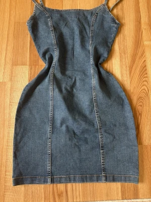 Jeansklänning med smala axelband - Snygg jeansklänning i blå denim med smala axelband och figurnära passform. Klänningen är kort och har markerade sömmar framtill och baktill som ger en cool look. Perfekt för dig som gillar enkel men trendig stil.