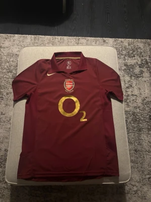 Arsenal Highbury tröja Henry 14 - Säljer en vinröd Arsenal Highbury jubileumströja från Nike med O2-logga i guld på framsidan och Henry 14 på ryggen. Tröjan har klassisk krage, broderat klubbmärke och detaljer i vitt och guld. Perfekt för dig som älskar fotboll och Arsenal.
