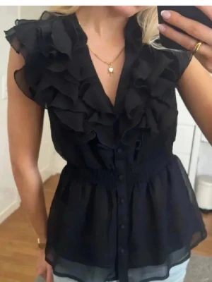 Svart volangblus med knappar - Snygg svart blus med volangdetaljer längs axlar och bröst, knappar framtill och markerad midja. Blusen är gjord i ett tunt, transparent tyg och har en feminin v-ringning. Perfekt för dig som gillar trendiga detaljer och en luftig känsla.