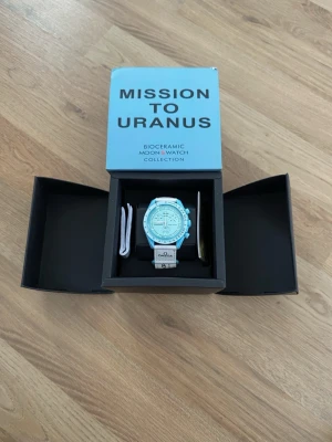 Omega x Swatch Mission to Uranus  - Säljer en Omega x Swatch Mission to Uranus från Bioceramic MoonSwatch Collection. Klockan har en ljusblå boett och vit rem med Omega och Swatch-logga. Snygg och unik design med kronograf och detaljerad urtavla. Perfekt för dig som gillar statement-accessoarer.