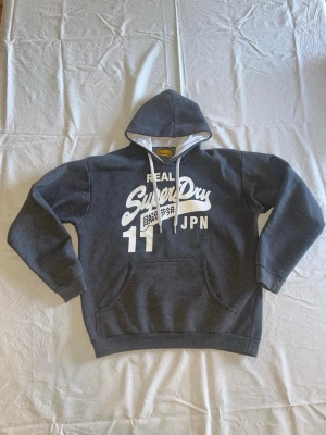 Mörkgrå Superdry hoodie med tryck - Mörkgrå hoodie från Superdry med vit logga och tryck framtill, stor ficka på magen och vita dragsnören i huvan. Klassisk streetstil med japanska tecken och siffran 11. Perfekt för en avslappnad look.