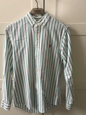 Randig skjorta från Polo Ralph Lauren - Snygg vit skjorta med gröna vertikala ränder från Polo Ralph Lauren. Slim fit-modell med klassisk krage och knäppning framtill. Broderad logga på bröstet. Tillverkad i bomull för en skön känsla och stilren look.