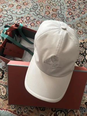 Loro Piana Keps - Snygg beige keps från Loro Piana med broderad logga framtill. Klassisk böjd skärm och justerbar resår baktill för perfekt passform. Tillverkad i ett slätt, exklusivt material som ger en clean och stilren look. Storlek XL motsvarar 60cm 