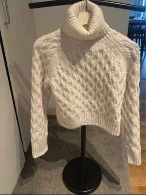 Vit kabelstickad polotröja - Säljer en vit kabelstickad polotröja med hög krage och långa ärmar. Tröjan har ett klassiskt flätmönster och ribbade muddar vid ärmslut och nederkant. Perfekt för kalla dagar och riktigt mysig att bära.