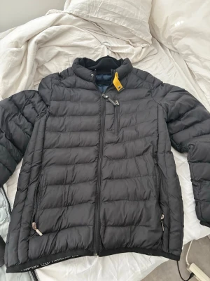 Svart pufferjacka från Parajumpers - Snygg svart pufferjacka från Parajumpers med gul detalj vid kragen och logga på axeln. Jackan har dragkedja framtill, två sidofickor med dragkedja och en bröstficka. Perfekt för kalla dagar tack vare det vadderade materialet. Passdar XL