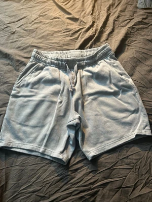 Ljusgråa mjuka shorts med snörning - Säljer ett par ljusgråa shorts med elastisk midja och snörning framtill. Shortsen har två sidofickor och en bakficka, tillverkade i ett mjukt frottéliknande material. Perfekta för chill dagar eller när du vill vara bekväm.