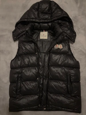 Svart dunväst från Moncler med huva - Snygg svart dunväst från Moncler med avtagbar huva och klassisk logotyp på bröstet. Västen har två fickor med dragkedja och är tillverkad i glansigt material med quiltad design. Perfekt för lager-på-lager och streetstyle.