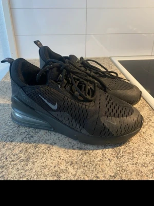 Nike Air Max 270 svarta sneakers - Snygga helsvarta Nike Air Max 270 sneakers med mesh-ovandel och synlig Air-enhet i hälen. Skorna har en sportig siluett, svarta skosnören och diskreta loggor på sidan och tungan. Perfekta för dig som gillar streetwear och vill ha bekväma sneakers med modern look. Storlek 38 men passar flera! Jätte sköna, kolla sista bild inte stort problem men ja