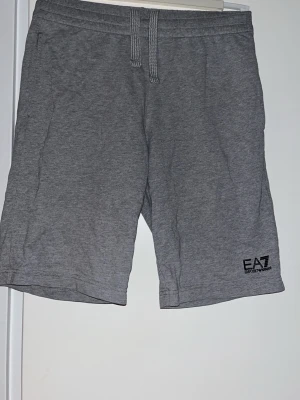Grå shorts från EA7 Emporio Armani - Snygga grå shorts från EA7 Emporio Armani med elastisk midja och snörning. Tillverkade i mjuk bomull för skön komfort. EA7-logga tryckt på vänster ben och diskreta ränder bak. Perfekta för chill eller träning.