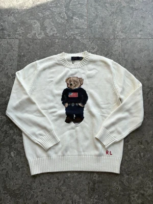 Vit stickad tröja Polo Bear - Vit stickad tröja från Polo Ralph Lauren med ikoniskt Polo Bear-motiv på bröstet. Björnen har en mörkblå tröja med amerikansk flagga och jeans. Rund halsringning och ribbade muddar. RL-broderi nertill. Viktigt att veta, den är liten i st, så den passar även dom med M!