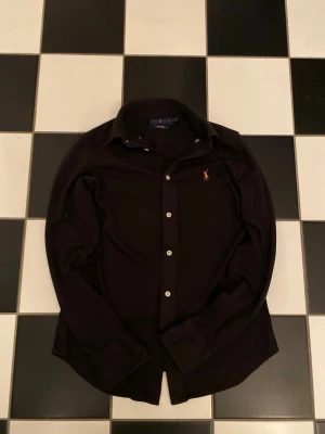 Svart skjorta från Ralph Lauren - Svart långärmad skjorta från Ralph Lauren, modell Knit Oxford. Klassisk krage, vita knappar och broderad logga på bröstet. Skjortan är gjord i bomull och har en normal passform som funkar till många olika stilar.