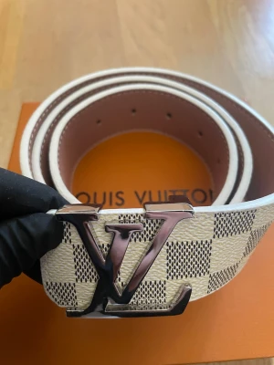Vit rutig bälte med metallspänne - Snyggt bälte i vitt och beige med rutigt mönster och ett stort spänne i metall. Perfekt accessoar för att lyfta din outfit med en exklusiv touch. Klassisk design som sticker ut.