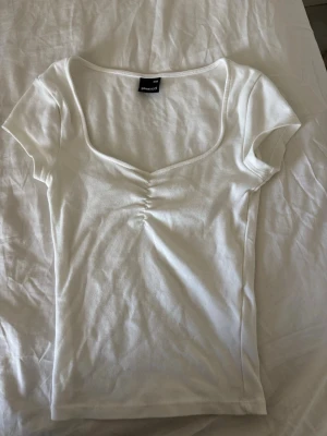 Vit topp med v-ringning från Gina Tricot - Snygg vit topp från Gina Tricot med v-ringning och lätt rynkdetalj framtill. Kortärmad och i mjukt bomullsmaterial, perfekt för en enkel och fräsch look. Passar till allt och är superenkel att styla.