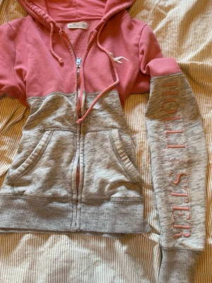 Rosa och grå hoodie från Hollister - Snygg hoodie från Hollister i tvåfärgad design med rosa upptill och grå nedtill. Dragkedja framtill, huva med dragsko och stora fickor. Hollister-logga broderad på ena ärmen och liten logga på bröstet. Perfekt för en chill och avslappnad stil.