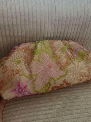 Broderad kuvertväska med blommor - Säljer en unik kuvertväska i beige tyg med broderade blommor i rosa, orange, grönt och lila. Väskan har paljettdetaljer och stängs med magnetknapp. Perfekt för dig som gillar färg och mönster. Insidan är ljus och har ett mindre fack.