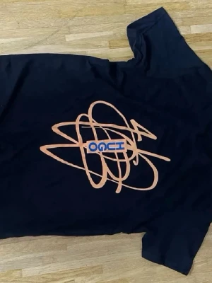 Svart t-shirt med orange grafiskt tryck - Cool svart t-shirt från OACI med ett stort orange grafiskt tryck på framsidan och blå textdetalj. Klassisk passform och rund halsringning. Perfekt för dig som gillar streetwear och unika prints.