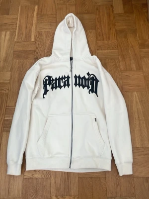Vit zip hoodie från Black Squad - Vit hoodie med dragkedja från Black Squad. Stor svart text i gotisk stil över bröstet. Mjuk insida och stor huva. Praktiska fickor framtill och ribbade muddar vid ärmslut och nederkant. I princip oanvänd. 