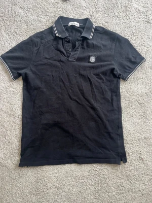 Svart pikétröja från Stone Island - Svart pikétröja från Stone Island med klassisk krage och knappar framtill. Tröjan har vit kontrastkant på krage och ärmslut samt en broderad logotyp på bröstet. Perfekt för en stilren och avslappnad look.