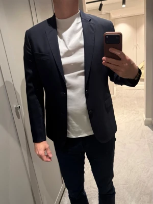 Marinblå kostym från J.Lindeberg  -  Säljer en stilren marinblå kostym med slimmad passform och snygga detaljer som knappar på ärmsluten och två framfickor. Kavajen har en bröstficka och är tillverkad i ett slätt material som ger ett elegant intryck. Perfekt för dig som vill ha en snygg o tidlös look. Modell: Hopper.