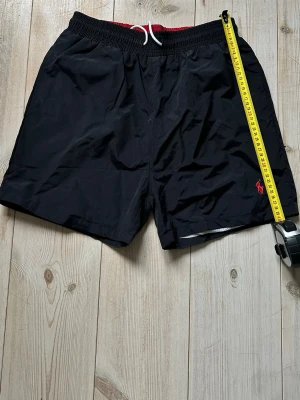 Svarta badbyxor från Polo Ralph Lauren - Snygga svarta badbyxor från Polo Ralph Lauren med röd insida och vit dragsko i midjan. Klassisk passform med elastisk midja och meshfoder. Liten röd logga broderad på benet. Perfekta för stranden eller poolen. Storlek M men väldigt liten i storleken. Helt nya och har inga defekter! Priset kan diskuteras!